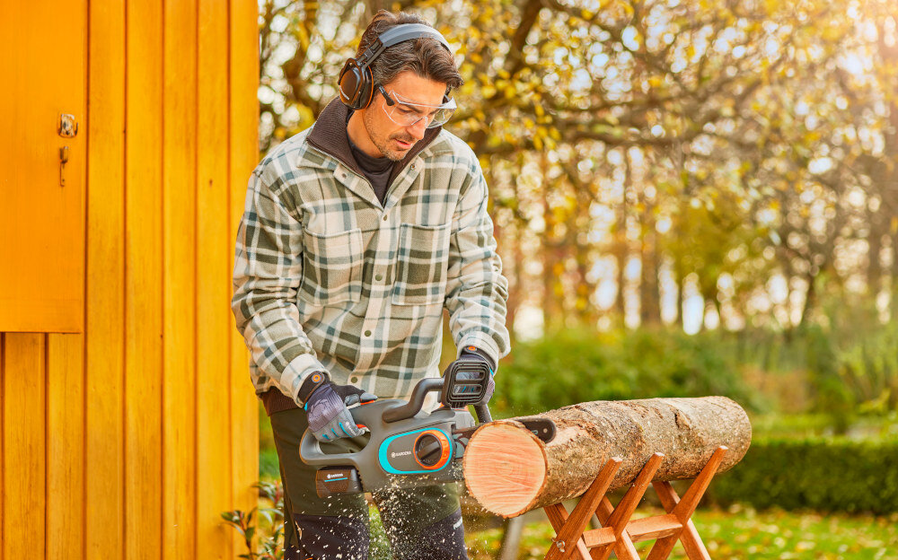 Piła akumulatorowa GARDENA PowerSaw 14790-55 osoba tnie kłodę drewna w tle drewniana ściana i drzewa bezszczotkowy silnik BLDC wysoka moc przy jednoczesnym niskim zużyciu energii maksymalna prędkość łańcucha sięga do 8 m/s mniej koniecznych konserwacji