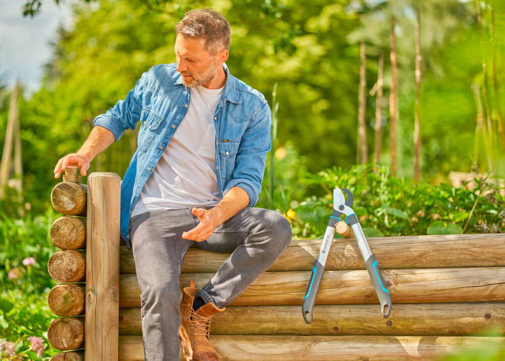 Nożyce do gałęzi GARDENA Easycut S 12030-20 ogród drzewa osoba siedzi na drewnianym płocie obok stoją nożyce oparte o płot długość 500 mm odpowiedni zasięg przy pracy wśród gęstych gałęzi dwuostrzowa konstrukcja uchwyt ErgoTec lekka aluminiowa budowa