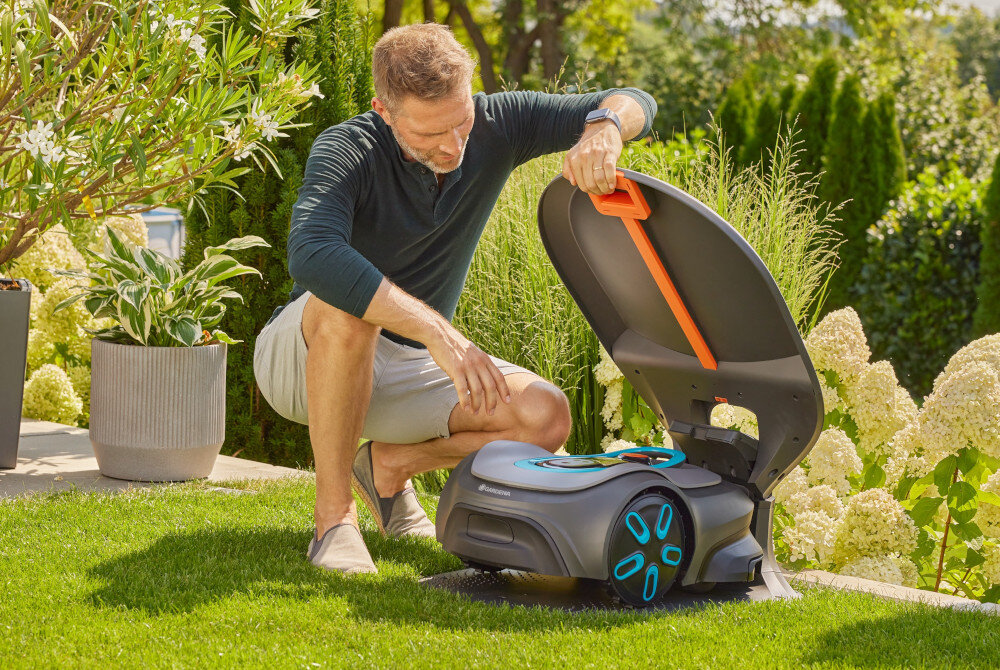 Garaż dla robota koszącego GARDENA Sileno Max Free 15025-20 ogród trawnik osoba podnosi klapę garażu w górę w garażu stoi robot koszący w tle kwiaty krzewy szybki dostęp do robota koszącego i jego elementów sterujących konstrukcja podnoszona do góry