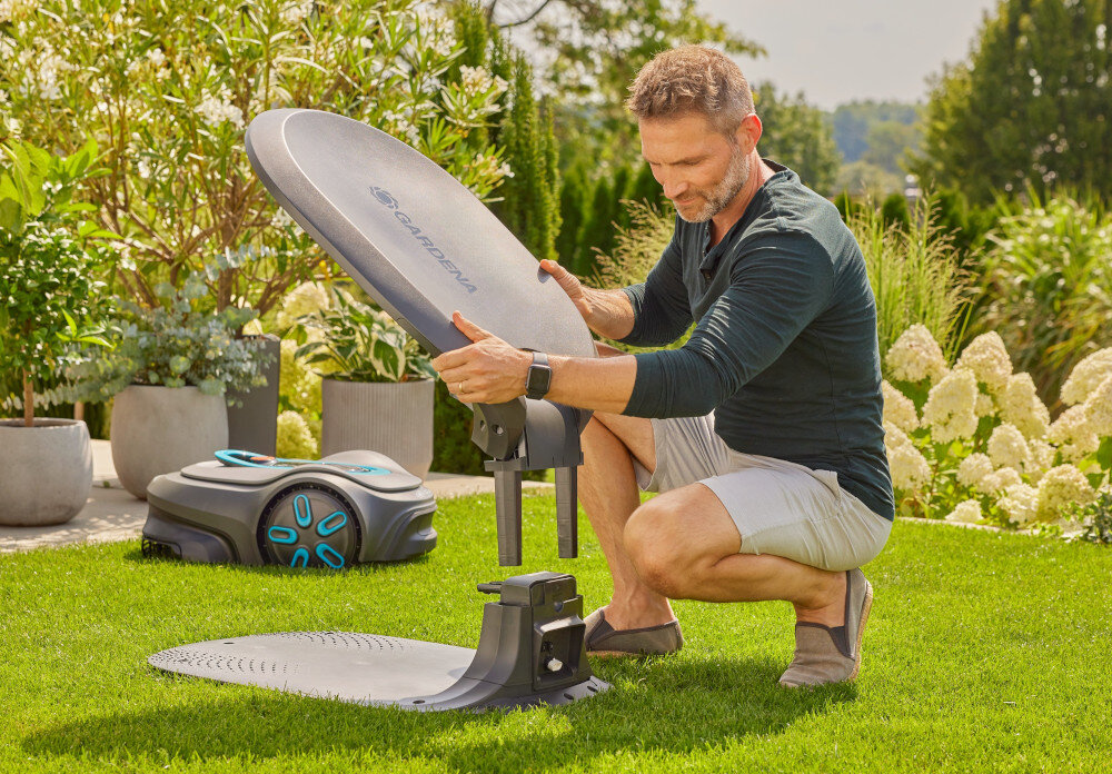 Garaż dla robota koszącego GARDENA Sileno Max Free 15025-20 ogród trawnik osoba montuje garaż na stacji ładującej obok stoi robot koszący w tle krzewy kwiaty w doniczkach montaż garażu nie wymaga użycia narządzi dopasowany do stacji ładującej robota SILENO z wytrzymałych materiałów tworzywo sztuczne odporny na deszcz grad i promieniowanie słoneczne w pełni funkcjonalne i bezpieczne w każdych warunkach