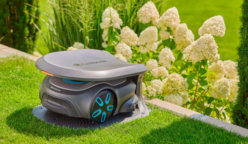 Garaż dla robota koszącego GARDENA Sileno Max Free 15025-20 ogród trawnik robot koszący umieszczony w garażu w tle kwiaty wymiary 38,2 x 70,7 x 52,1 cm waga około 4,9 kg ułatwia przenoszenie i ewentualną zmianę lokalizacji