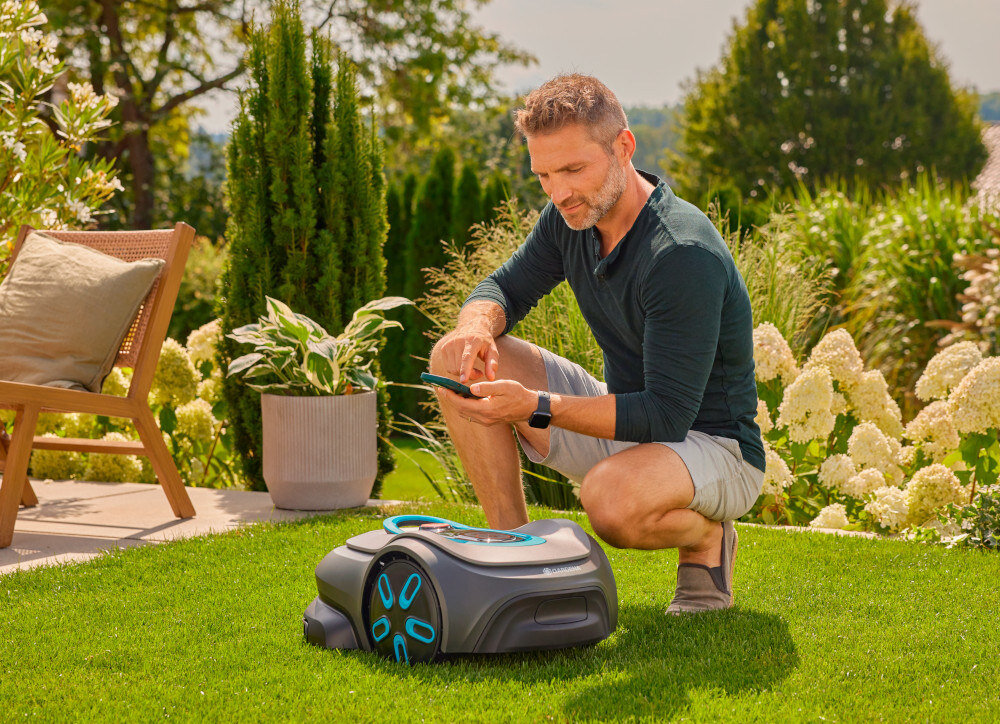 Robot koszący GARDENA Sielno Max 1500 19903-32 robot stoi na trawie osoba kuca obok z telefonem w dłoni tworzenie harmonogramu koszenia aktualizacje realizowane bezprzewodowo dzięki funkcji FOTA moduł Bluetooth łaczność z aplikacją GARDENA Bluetooth® App lub GARDENA Smart App na systemy iOS 17/Android 9 lub nowsze