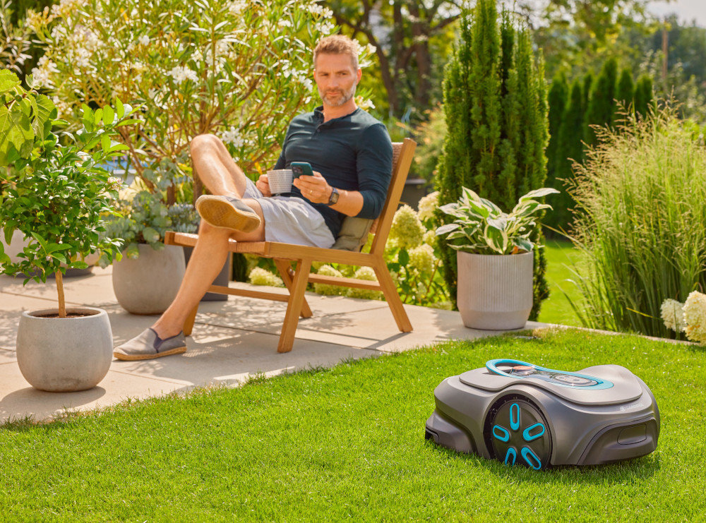 Robot koszący GARDENA Sielno Max 1500 19903-32 robot stoi na trawie w tle osoba siedzi na krześle na tarsie z telefonem w dłoni poziom hałasu 58 dB(A) akumulator litowo-jonowy o napięciu 18 V i pojemności 2,0 Ah dzienny czas pracy do 22 godzin