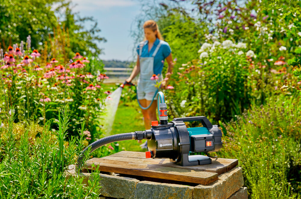 Pompa ogrodowa GARDENA 3700 Basic 9014-47 elektryczna pompa ustawiona na drewnianej podłodze w tle osoba podlewa rośliny w ogrodzie uniwersalne zastosowanie do wody czystej i lekko zabrudzonej do pompowania wody źródło zasilania zraszaczy wydajny system nawadniania
