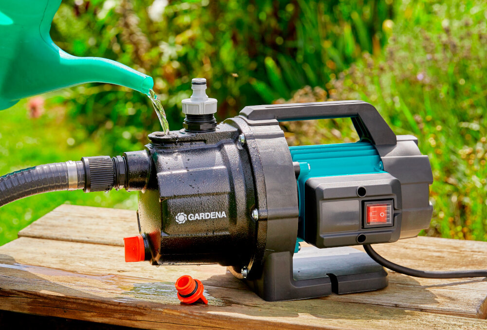 Pompa ogrodowa GARDENA 3700 Basic 9014-47 elektryczna pompa ustawiona na drewnianej podłodze osoba napełnia pompę wodą z konewki w tle rośliny duży otwór wlewowy szybkie i wygodne napełnianie śruba spustowa sprawne opróżnienie resztek cieczy ochrona przed uszkodzeniem w wyniku zamarznięcia wody wewnątrz obudowy