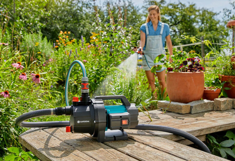 Pompa ogrodowa GARDENA 3000 Basic 09010-47 elektryczna pompa ustawiona na drewnianej podłodze w tle osoba podlewa rośliny w ogrodzie uniwersalne zastosowanie do wody czystej i brudnej do pompowania wody źródło zasilania zraszaczy wydajny system nawadniania