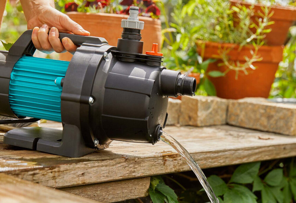 Pompa ogrodowa GARDENA 3000 Basic 09010-47 elektryczna osoba trzyma pompę za uchwyt opróżnia pompę z wody w tle rośliny praktyczny zintegrowany uchwyt ułatwia przenoszenie waga 5,3 kg kabel zasilający o długości 1,5 metra