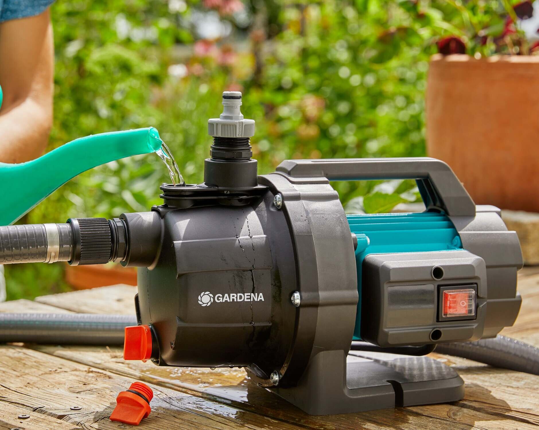 Pompa ogrodowa GARDENA 3000 Basic 09010-47 elektryczna pompa ustawiona na drewnianej podłodze osoba napełnia pompę wodą z konewki w tle rośliny duży otwór wlewowy szybkie i wygodne napełnianie śruba spustowa sprawne opróżnienie resztek cieczy ochrona przed uszkodzeniem w wyniku zamarznięcia wody wewnątrz obudowy