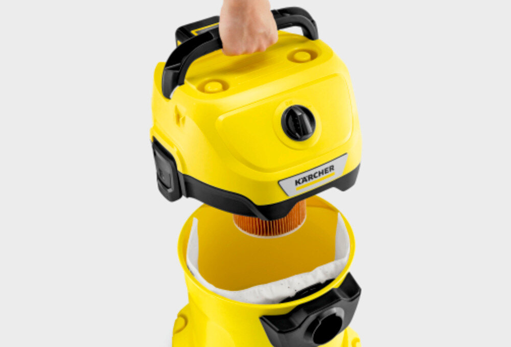 Żółty odkurzacz z logo \'KARCHER\' na górnej części, która jest podnoszona ręką. Wewnątrz widoczny jest pomarańczowy filtr i biały worek.