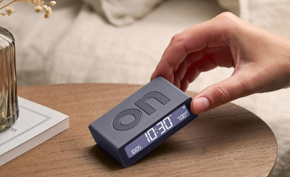 Budzik LEXON Flip Mini Ciemnoszary, ergonomia, ON, OFF, obrót, Ręka przechyla mały budzik Lexon Flip Mini w kolorze grafitowym, stół, książka, neutralne tło.