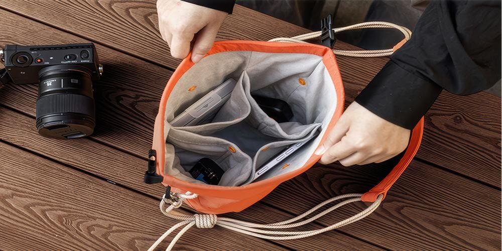 Otwarta pomarańczowa torba z jasnymi przegrodami na drewnianym blacie; dłonie trzymają krawędzie, obok leży aparat