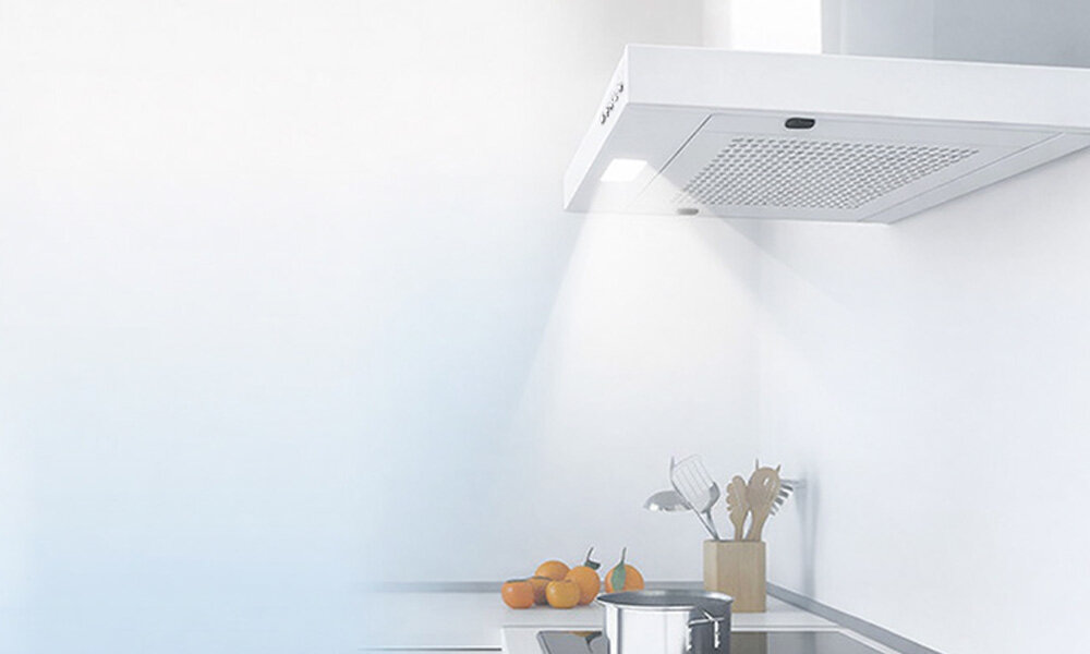 OKAP MIDEA MH60F17EM21WB Ilustracja przedstawia oświetlony okap kuchenny emitujący światło na płytę grzewczą poniżej. W tle widać jasną, minimalistyczną kuchnię z owocami i przyborami kuchennymi oświetlenie LED Energooszczędne klasa B jasne równomierne światło