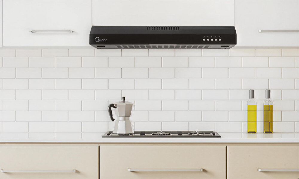 OKAP MIDEA MH60F17EM21BB Na zdjęciu widać czarny okap kuchenny zamontowany nad płytą gazową. Przed nim stoi dzbanek do parzenia kawy oraz dwie butelki z oliwą, tworząc estetyczny, minimalistyczny wystrój kuchni