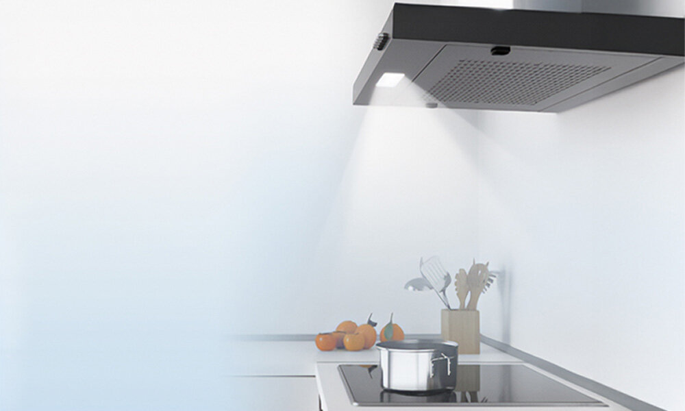 OKAP MIDEA MH60F17EM21BB Ilustracja przedstawia oświetlony okap kuchenny emitujący światło na płytę grzewczą poniżej. W tle widać jasną, minimalistyczną kuchnię z owocami i przyborami kuchennymi oświetlenie LED Energooszczędne klasa B jasne równomierne światło