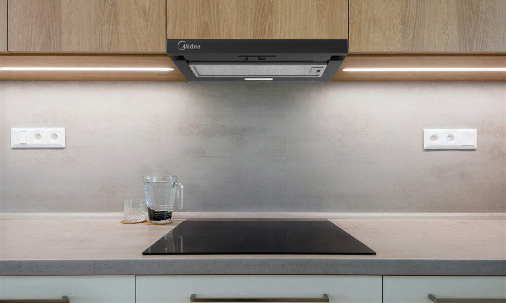 OKAP MIDEA MH60L07EM22BB Fotografia ukazuje kuchnię z okapem zabudowanym w szafce wiszącej. Pod okapem znajduje się płyta indukcyjna na czystym, minimalistycznym blacie.