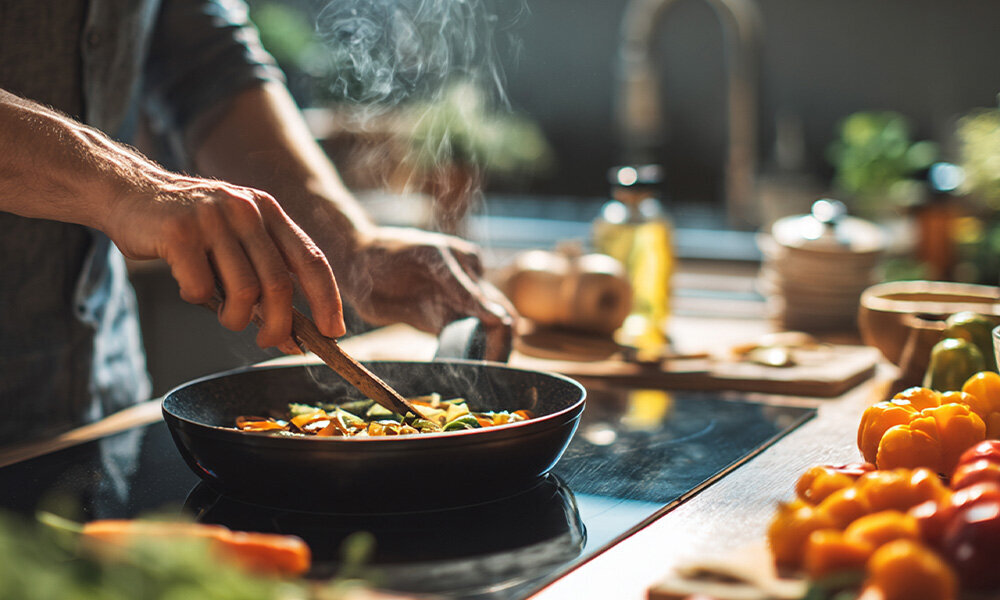 OKAP MIDEA MH60L07EM22BB Na zdjęciu widać osobę przygotowującą posiłek na patelni na płycie kuchennej. Kolorowe warzywa oraz para unosząca się znad patelni tworzą atmosferę gotowania. moc wydajność cicha praca 65 dB