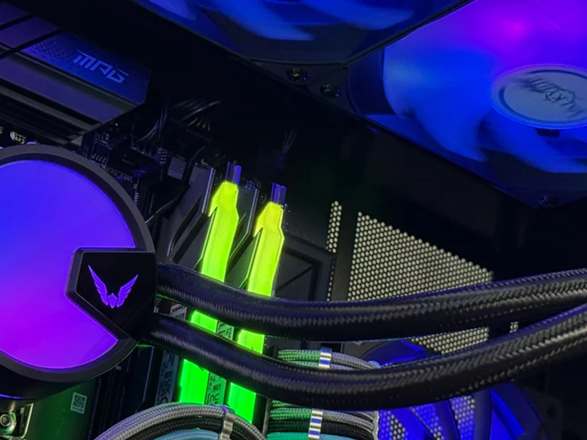 Obraz przedstawia wnętrze komputera z zamontowanym chłodzeniem wodnym AIO, które ma podświetlenie RGB w kolorach fioletowym i niebieskim. Widoczne są również dwa podświetlone na zielono moduły pamięci RAM oraz wentylator z niebieskim podświetleniem.