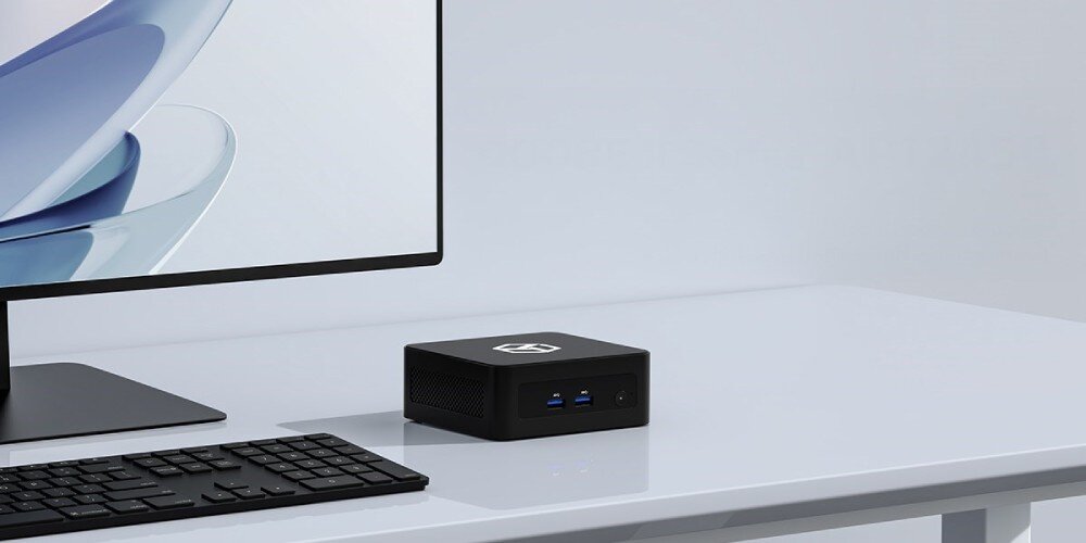 Komputer QOOBE II AP-1340 Małe czarne urządzenie z logo na górze stoi na białym biurku obok monitora, klawiatury i myszki, w tle widać fragment tapety z zaokrąglonym kształtem, opis