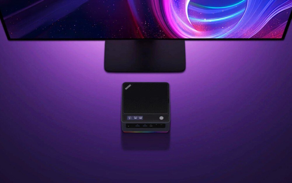Komputer QOOBE U155H Widok z góry na mini pc z wyświetlaczem na obudowie, ustawiony na biurku pod monitorem na fioletowym tle, urządzenie z podświetleniem led u podstawy, karta graficzna, obraz
