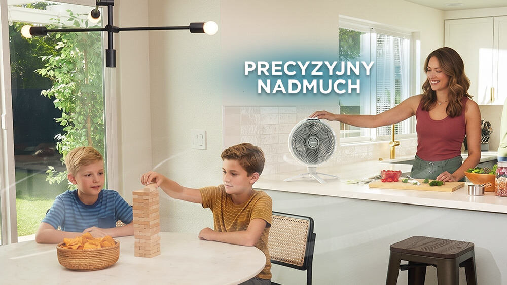Kobieta w kuchni obsługuje wentylator stojący na blacie, obok niej znajdują się pokrojone owoce. Na pierwszym planie dwóch chłopców gra w grę z drewnianymi klockami, a nad nimi widnieje napis \'PRECYZYJNY NADMUCH\'.