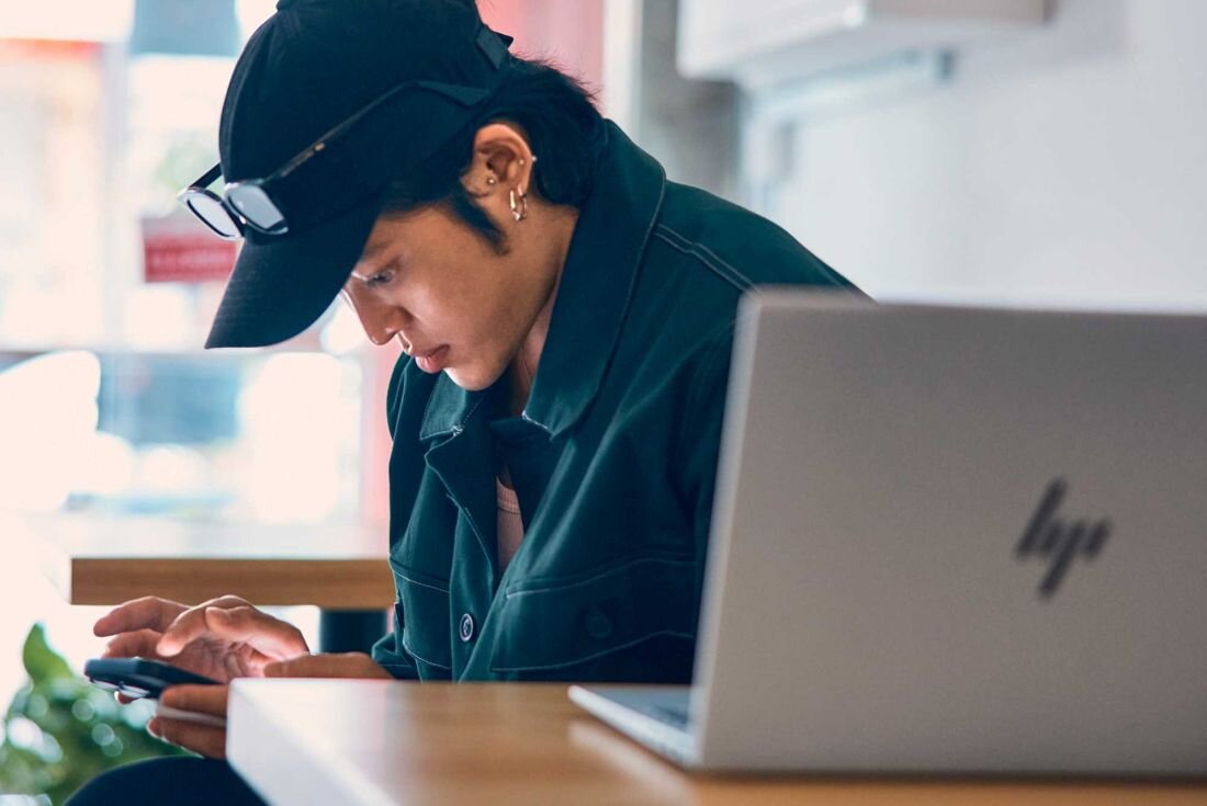 Mężczyzna w czapce baseballowej patrzy na swój telefon, siedząc obok laptopa HP Pavilion Plus 14. Pamięć RAM LPDDR5X, Dysk SSD PCIe NVMe