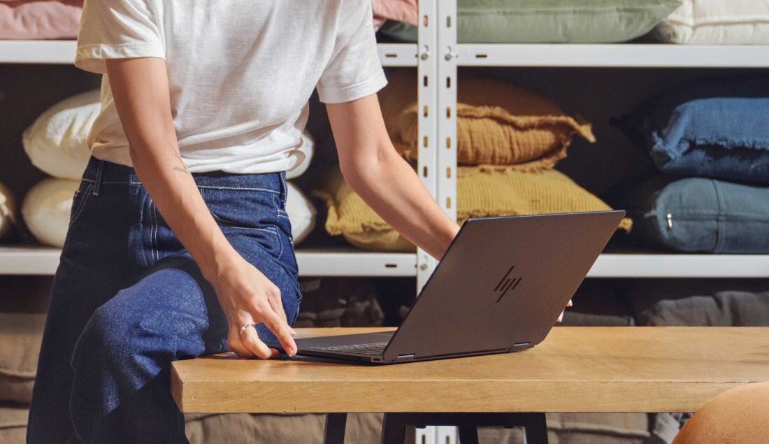 Kobieta siedzi przy drewnianym stole, korzystając z laptopa HP OmniBook Ultra Flip. baterię o pojemności 64 Wh