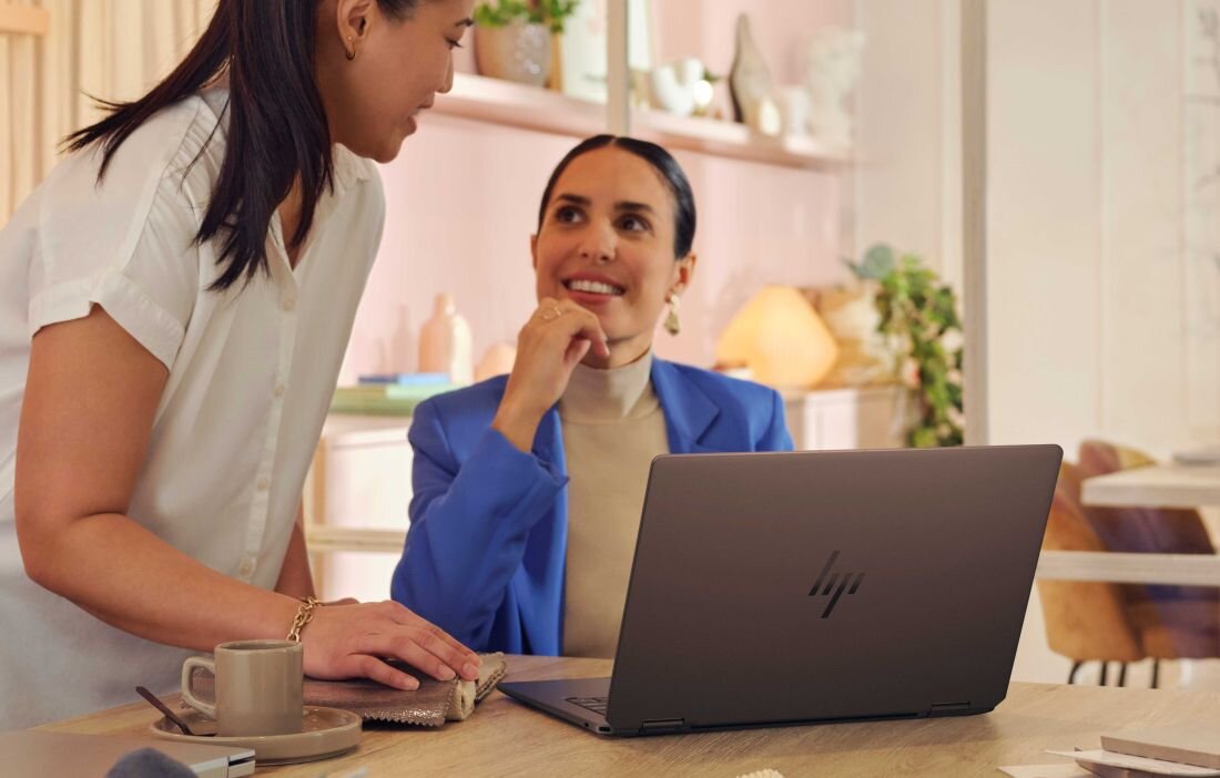 Dwie kobiety rozmawiają przy laptopie HP OmniBook Ultra Flip, siedząc przy biurku. Pamięć wewnętrzna LPDDR5X