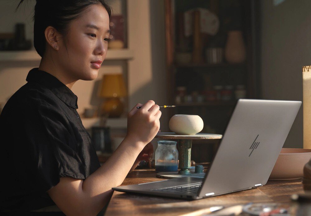 Laptop HP Envy x360 14-FA0014NW Kobieta w czarnej koszuli siedzi przed laptopem, trzymając pędzel w dłoni i patrząc na niewielką glinianą rzeźbę na stole, opis