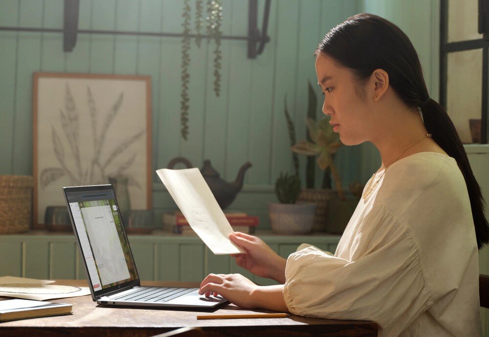 Laptop HP Envy x360 14-FA0014NW Kobieta w jasnej bluzce siedzi przy stole, patrząc na ekran laptopa i trzymając w dłoni kartkę papieru, procesor, rdzenie, pamięć RAM