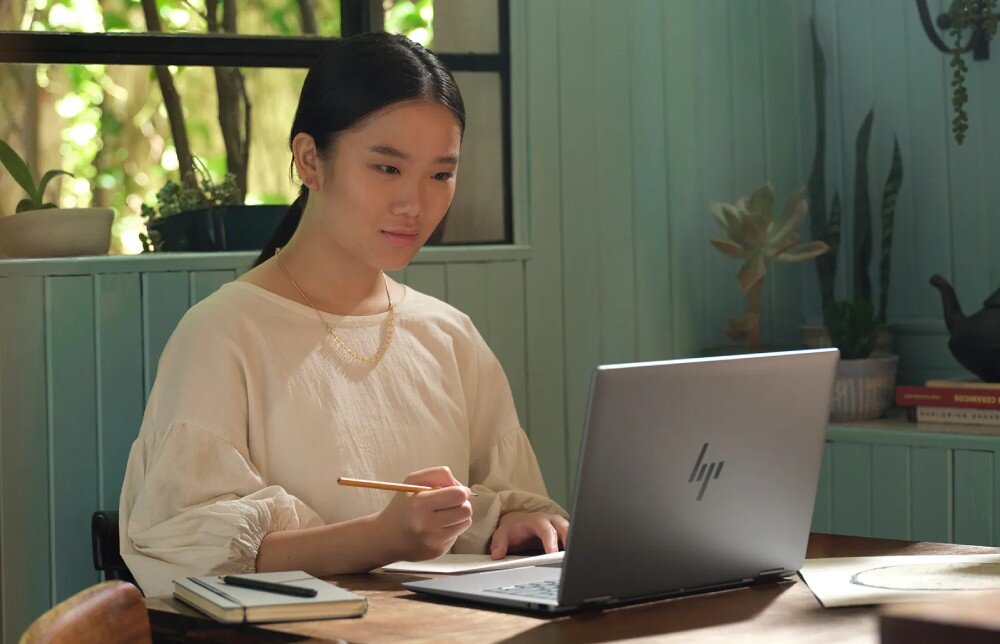 Laptop HP Envy x360 14-FA0014NW Kobieta siedząca przy biurku w jasnym, nasłonecznionym pomieszczeniu używa laptopa, trzymając ołówek, kamera internetowa, mikrofony, głośniki