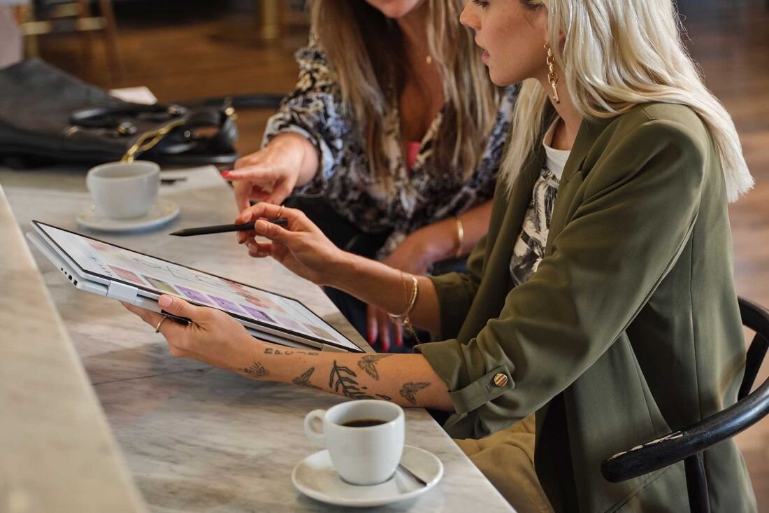 Dwie kobiety siedzą przy stole z laptopem HP Envy x360, rozmawiając i pracując nad projektem. bateria litowo-polimerową (LiPo) 68 Wh z czterema komorami   