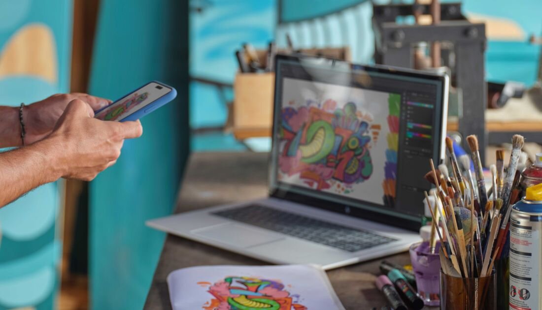 Mężczyzna korzysta z telefonu, siedząc przy stole z laptopem HP Envy x360 i zestawem przyborów artystycznych.  Grafika Intel wspierana przez AI Intel AI Boost   