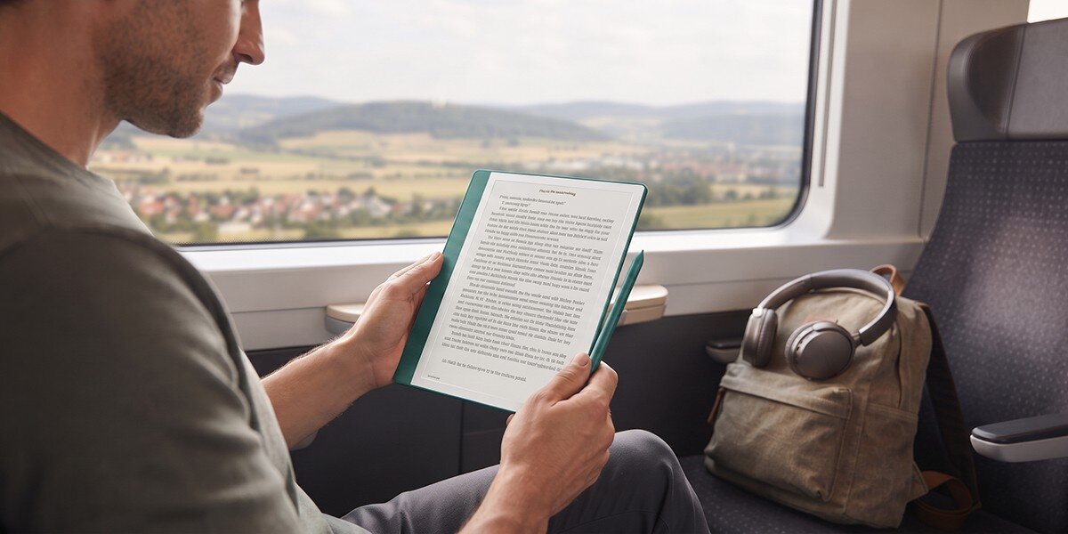 Osoba siedzi w pociągu i trzyma czytnik e-booków z wyświetlonym tekstem. Obok na siedzeniu znajduje się plecak z słuchawkami.