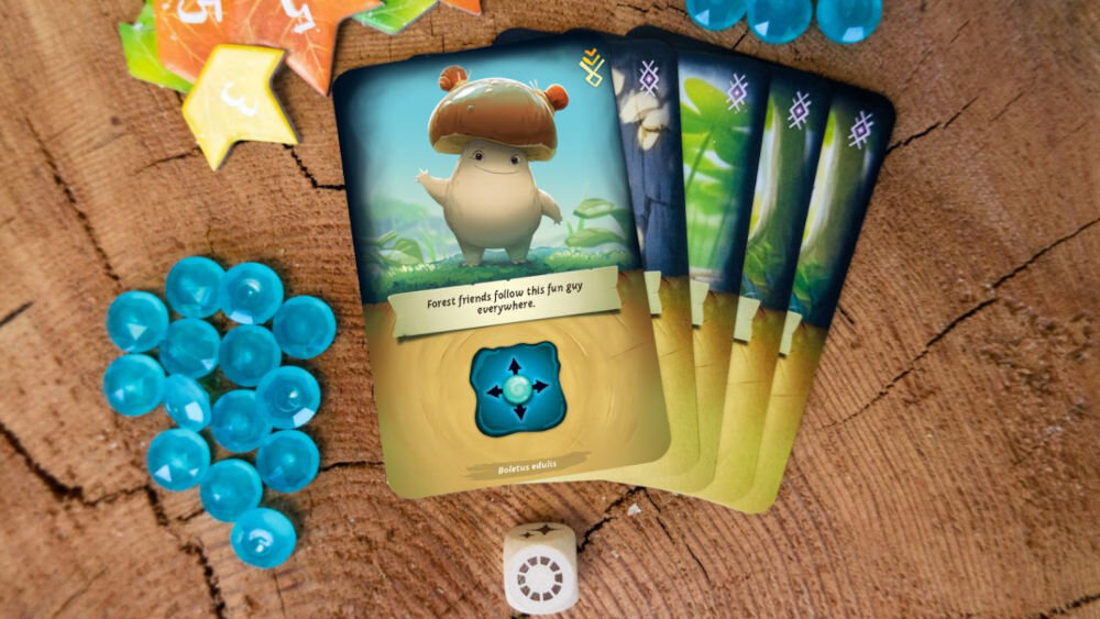 Gra planszowa RAVENSBURGER Mycelia 22898 karty rozłożone na pienku niebieskie kulki mechanika gry budowanie talii ilustracje inspirowane grzybami gra strategiczne dla rodziny dla przyjaciół