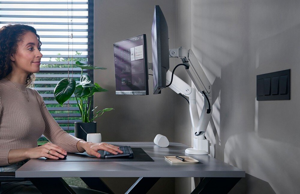 Uchwyt do monitora ONE FOR ALL DM4220 Kobieta siedzi przy biurku i pracuje na dwóch monitorach zamocowanych na białym uchwycie z podwójnym ramieniem, opis