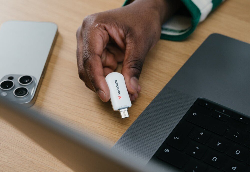 Pendrive VERBATIM Plectra Dłonie umieszczające czarny pendrive z portem USB-C na drewnianym blacie obok laptopa i smartfona