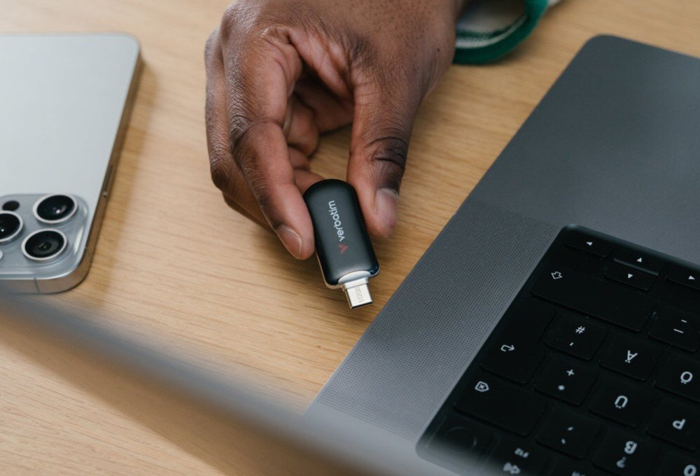 Pendrive VERBATIM Plectra Dłonie umieszczające czarny pendrive z portem USB-C na drewnianym blacie obok laptopa i smartfona