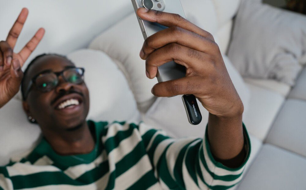 Pendrive VERBATIM Plectra Osoba leżąca na kanapie w okularach trzyma smartfon w jednej ręce oraz czarny pendrive w drugiej ręce