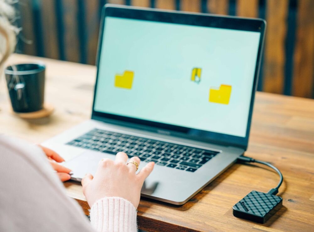 Osoba korzysta z laptopa, do którego podłączony jest zewnętrzny dysk SSD o wzorze przypominającym diament. Na ekranie laptopa widoczne są dwa żółte foldery.