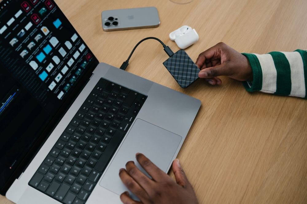 Osoba trzyma zewnętrzny dysk SSD podłączony do laptopa, na którym widać otwarte okno z ikonami. Obok leży para bezprzewodowych słuchawek i smartfon.
