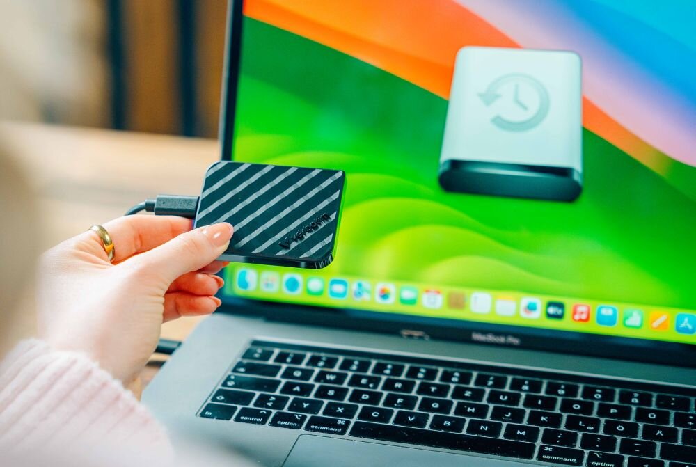 Osoba trzyma czarny dysk z napisem \'Verbatim\', podłączony do laptopa z kolorowym ekranem. Na ekranie widoczna jest ikona dysku z symbolem zegara.