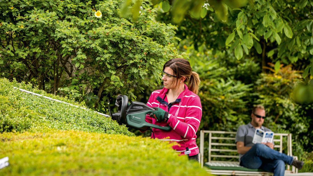 Nożyce do żywopłotu BOSCH AdvancedHedgeCut 70 0.600.8C0.905 ogród drzewa krzewy osoba przycina żywopłot w tle siedzi osoba na ławce czyta książkę dźwiękochłonna obudowa z technologią ProSilence obniża poziom hałasu o 80%, do 93 dB(A) komfort pracy stosowanie nożyc w miejscach blisko domów lub osiedli sprzęgło ślizgowe o momencie obrotowym 50–70 Nm cięcie grubszych gałęzi chroni silnik przed przeciążeniem płynne działanie narzędzia
