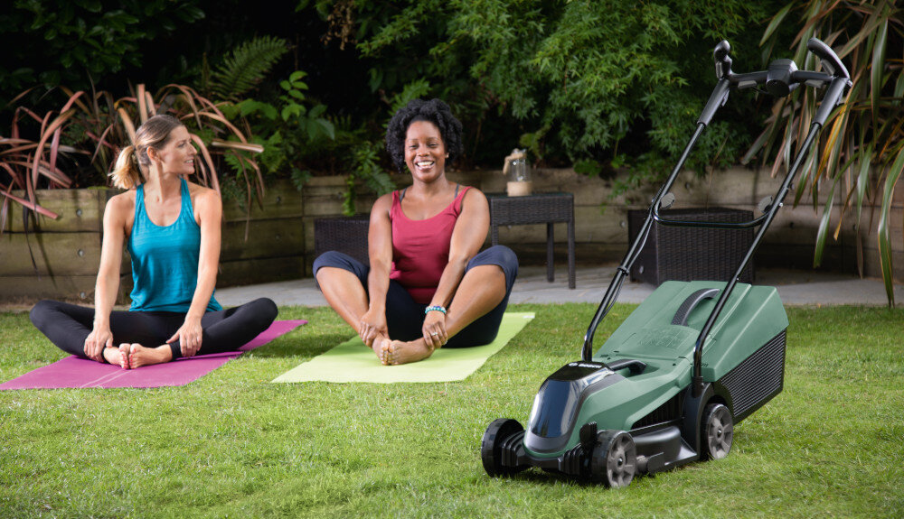 Kosiarka akumulatorowa BOSCH CityMower 0.600.8B9.A09 kosiarka na trawniku w tle dwie kobiety ćwiczą na matach ogród rośliny zadbany trawnik piękna trawa mobilność zasilanie akumulatorowe