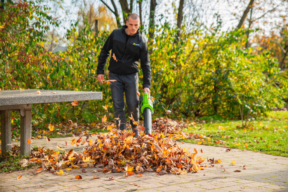 Dmuchawa do liści akumulatorowa 4GARDEN (bez akumulatora) AD261 podjazd chodnik osoba zdmuchuje liście z chodnika w tle drzewa krzewy niebo sprzątnie porządek wszechstronne zastosowanie