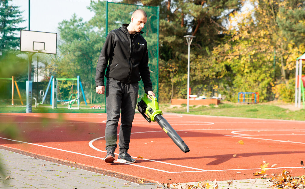 Dmuchawa do liści akumulatorowa 4GARDEN (bez akumulatora) AD261 boisko osoba zdmuchuje liście z boiska w tle niebo drzewa słup do koszykówki akumulatorowy system 4GARDEN 20 V i 40 V rozwiązanie systemowe 4GARDEN oferuje większą wygodę użytkowania i lepszą organizację pracy