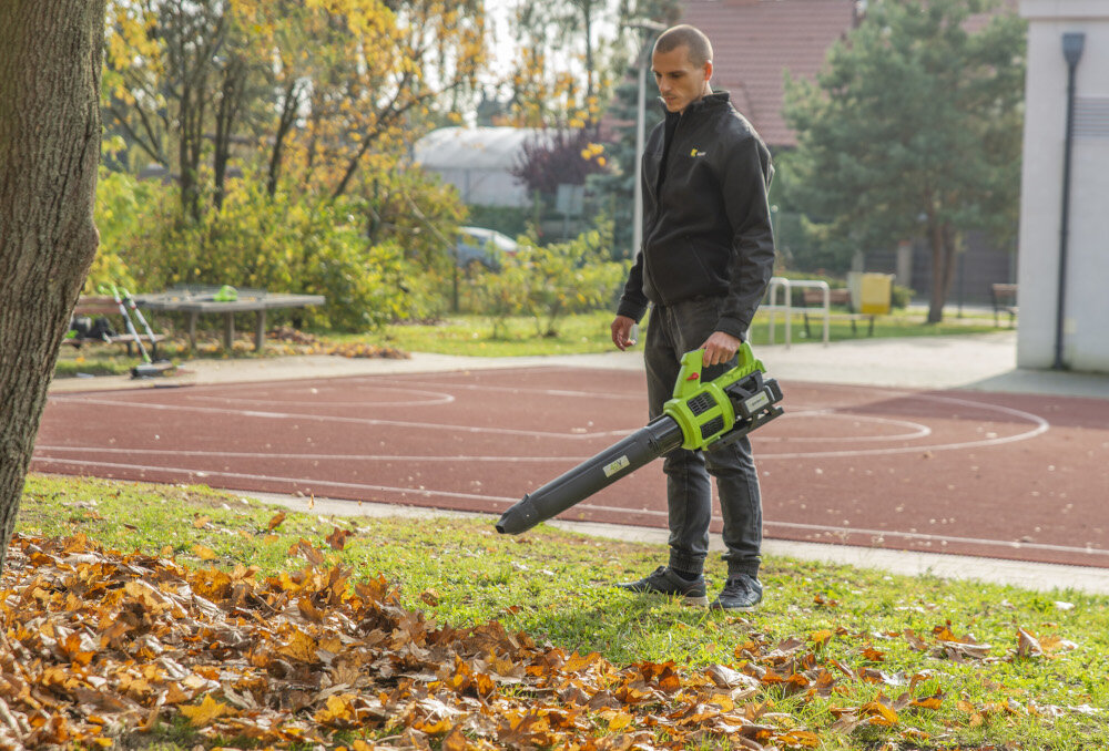 Dmuchawa do liści akumulatorowa 4GARDEN (bez akumulatora) AD261 boisko osoba zdmuchuje liście z boiska w tle drzewa krzewy budynek waga około 0,9 kg miękki uchwyt minimalizuje wibracje koła ułatwiają prowadzenie ergonomiczna budowa poziom hałasu do 102 dB silnik utrzymuje prędkość obrotową nawet przy dużym obciążeniu stała moc szybkie rezultaty