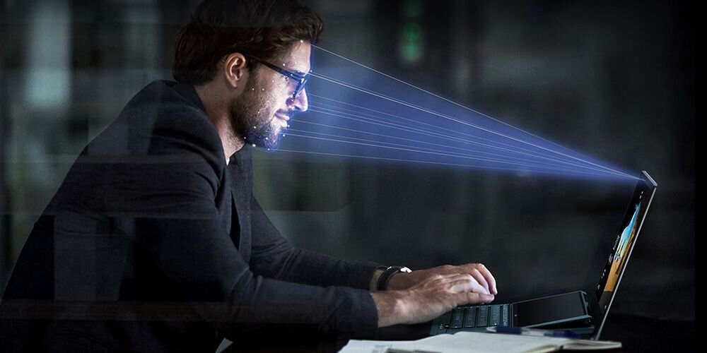 Mężczyzna pracujący przy laptopie ASUS Zenbook 14, wiązki światła skierowane na twarz – ilustracja działania kamery IR do rozpoznawania twarzy.