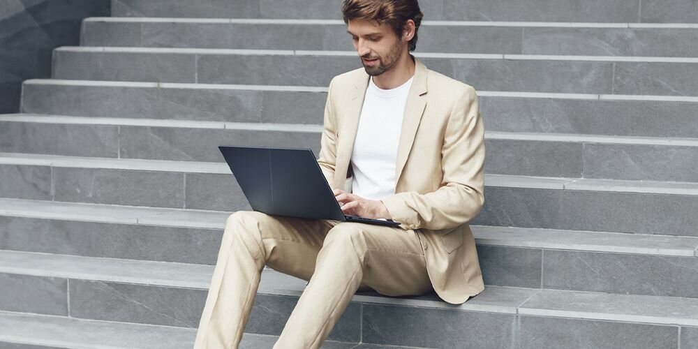 Mężczyzna w jasnym garniturze siedzący na schodach z laptopem ASUS Zenbook 14 – praca na ultrabooku z baterią 75 Wh, styl biznesowy.
