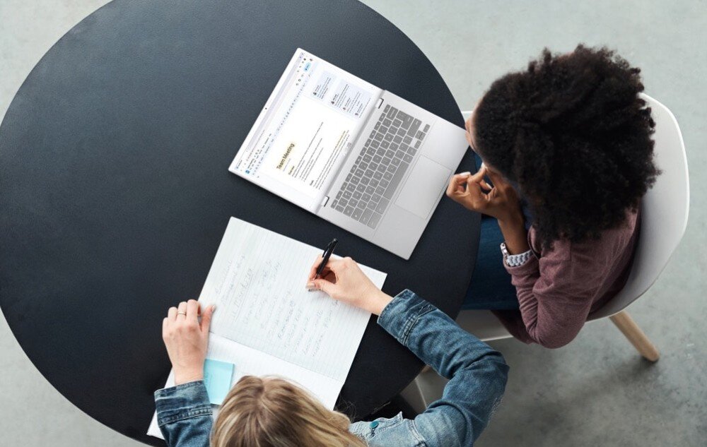 Laptop ASUS Chromebook Plus CB3402CVA-MW0529 Widok z góry na dwie kobiety przy okrągłym stole, współpracujące przy laptopie. Jedna kobieta robi notatki na papierze, druga patrzy na ekran komputera, wyświetlacz, rozdzielczość, matryca, odchylanie, zawias