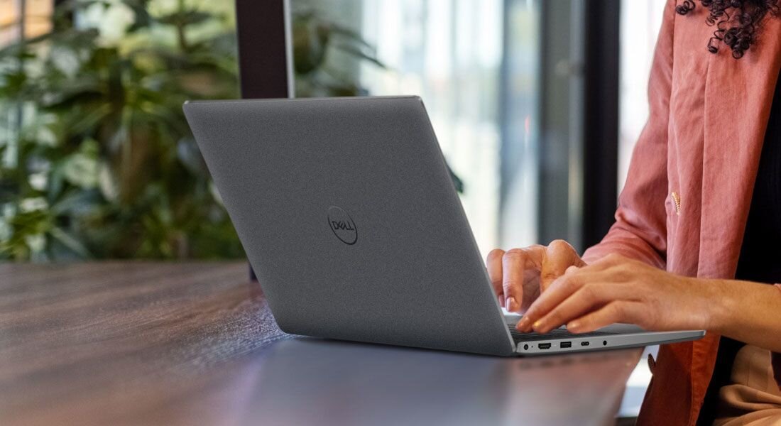 Zbliżenie na dłonie kobiety piszącej na klawiaturze laptopa DELL Latitude przy stole w loftowym wnętrzu z roślinami. - Opis serii Latitude 3450  